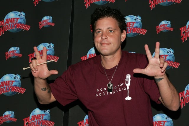 Corey Haim in a red tshirt, waving his hands in front of a Planet Hollywood wallpaper