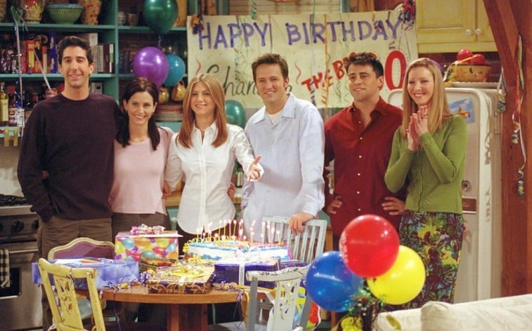 The cast of Friends stands around a table with a birthday cake