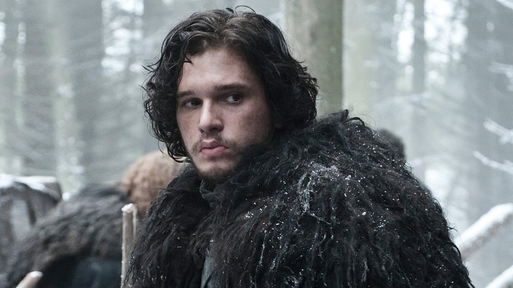 Jon Snow is in a black coat in the snow looking sad.