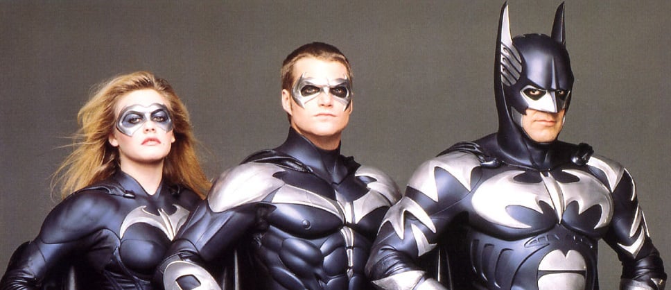 Batman and Robin stand in uniform looking into the camera in front of a grey background