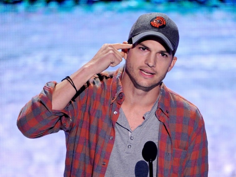 Ashton Kutcher in a Chicago Bears baseball cap and plaid shirt speaks into a microphone and points at his head