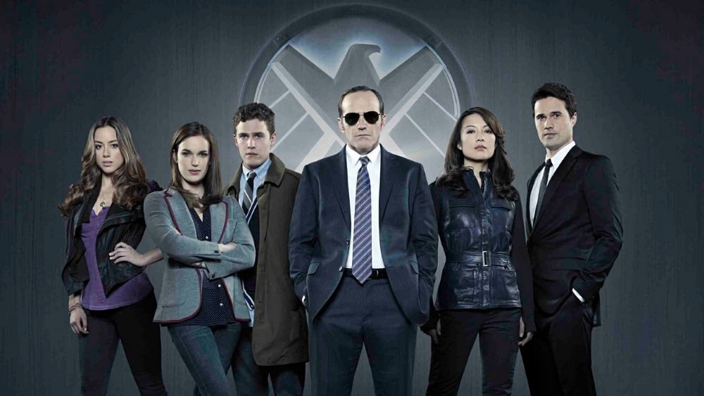Cast of Agents of SHIELD stand next to one another in front of the SHIELD emblem