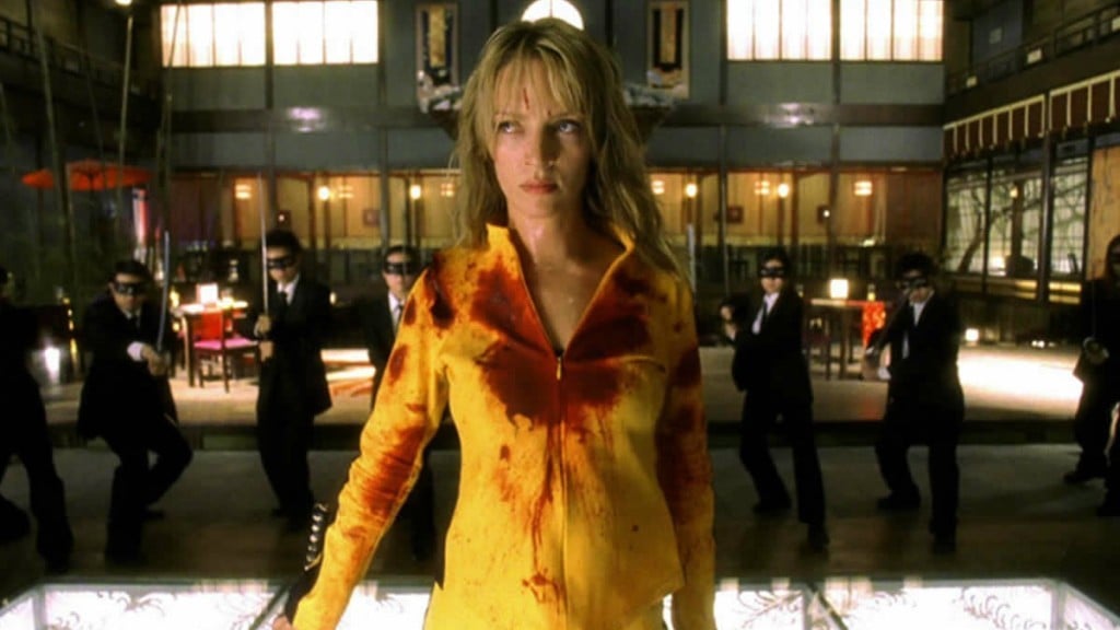 The Bride stands in a blood-covered yellow jump suit surrounded by men in suits.