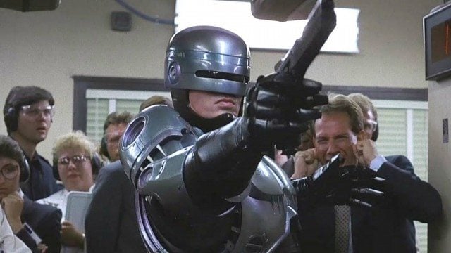 Robocop stands in front of a crowd in a hallway and is pointing a gun in the opposite direction.