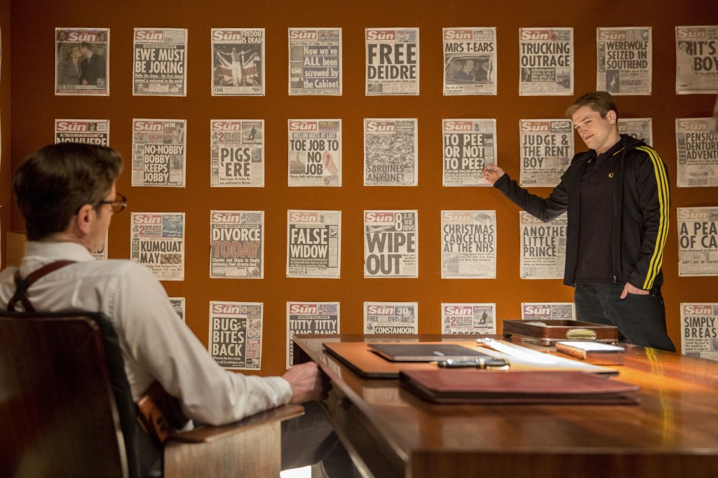 Taron Egerton in front of a wall of newspapers, pointing to one toward the middle
