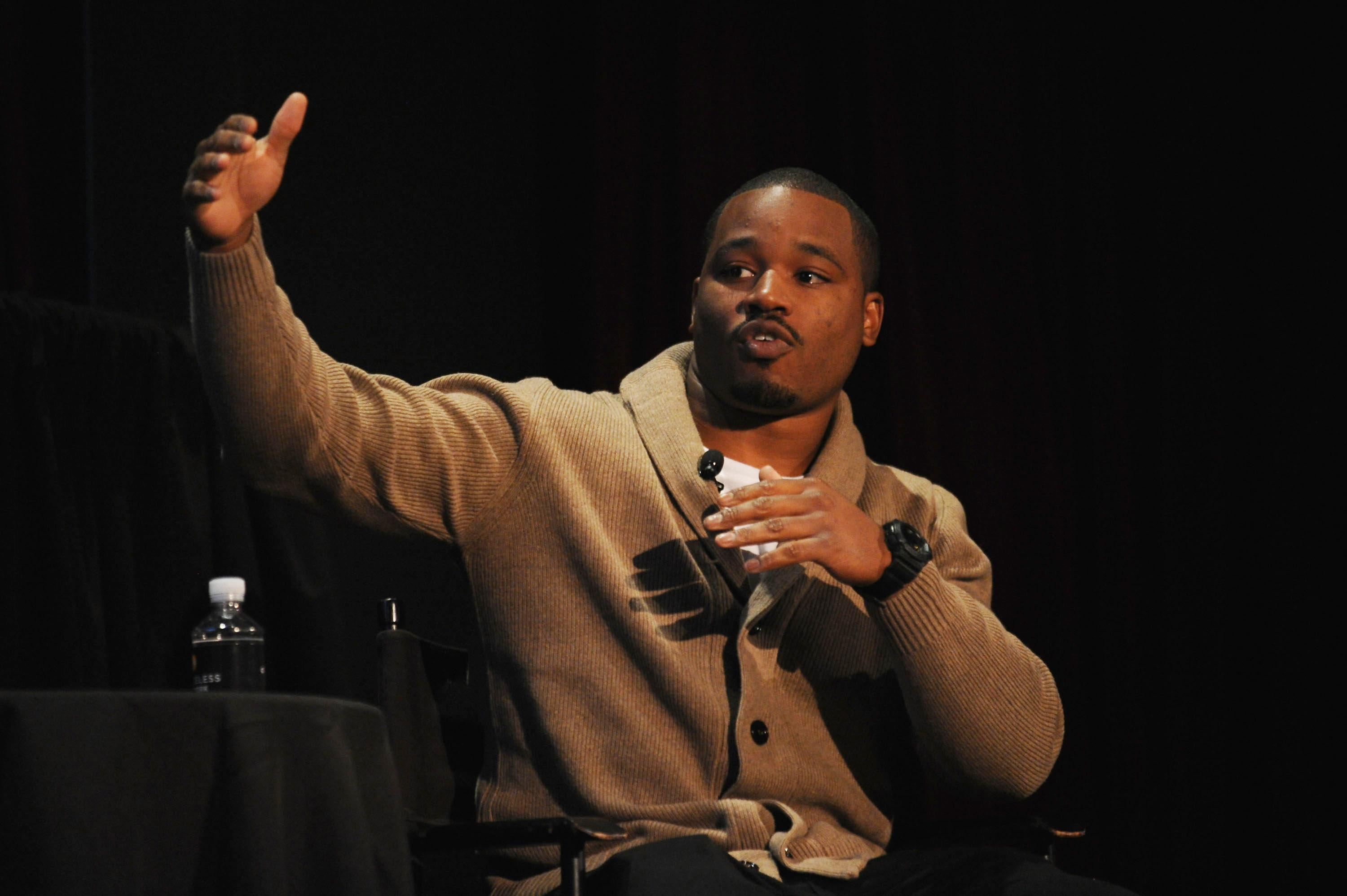 Ryan Coogler speaks at a forum, with his right arm extended up and out
