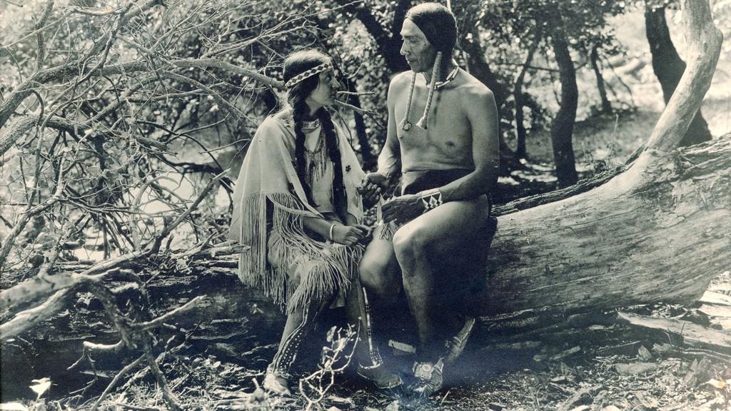 A Native American woman and man sitting on a fallen tree in the forest in a scene from The Daughter of Dawn