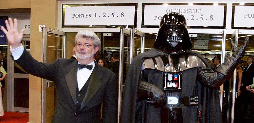 George Lucas in a tux waving next to a figurine of Darth Vader waving