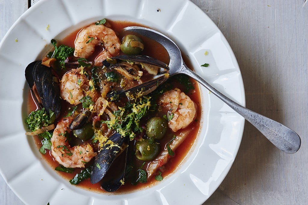 seafood stew with olives, mussels, shrimp, and broccoli rabe in a white bowl