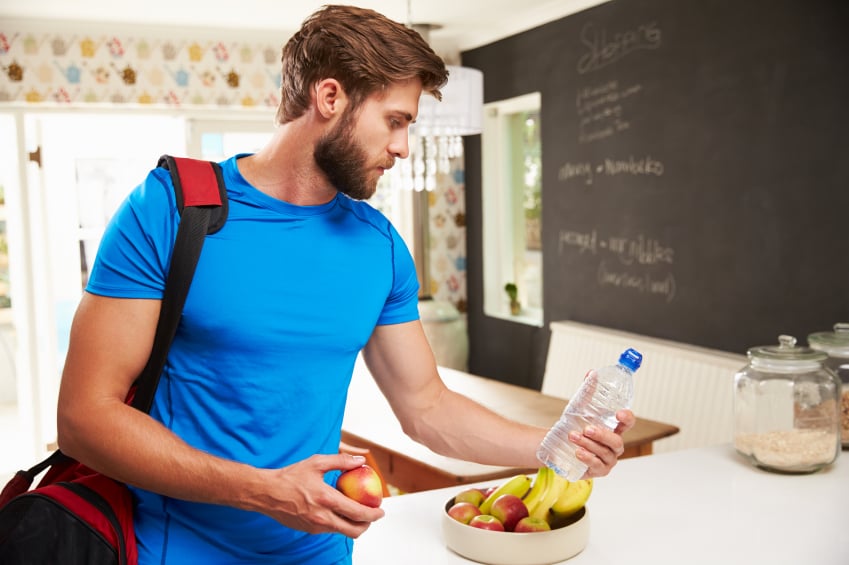 Man wearing gym clothes and a gym bag grabs fruit and a bottle of water for after his workout