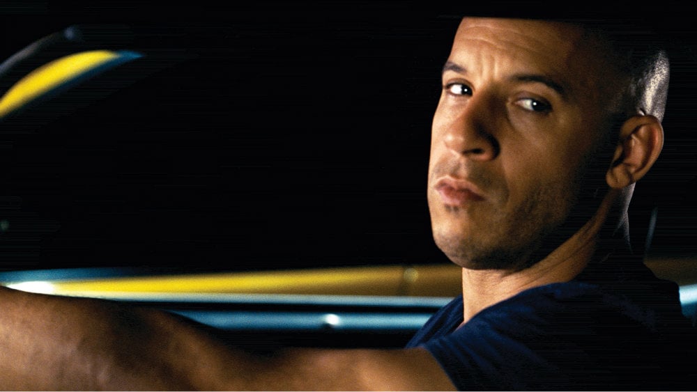 Vin Diesel in Fast Five is looking to the side in the driver's seat of a car.
