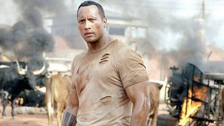 A sweat-caked Dwayne Johnson wearing a torn tshirt, looking to the left of the frame