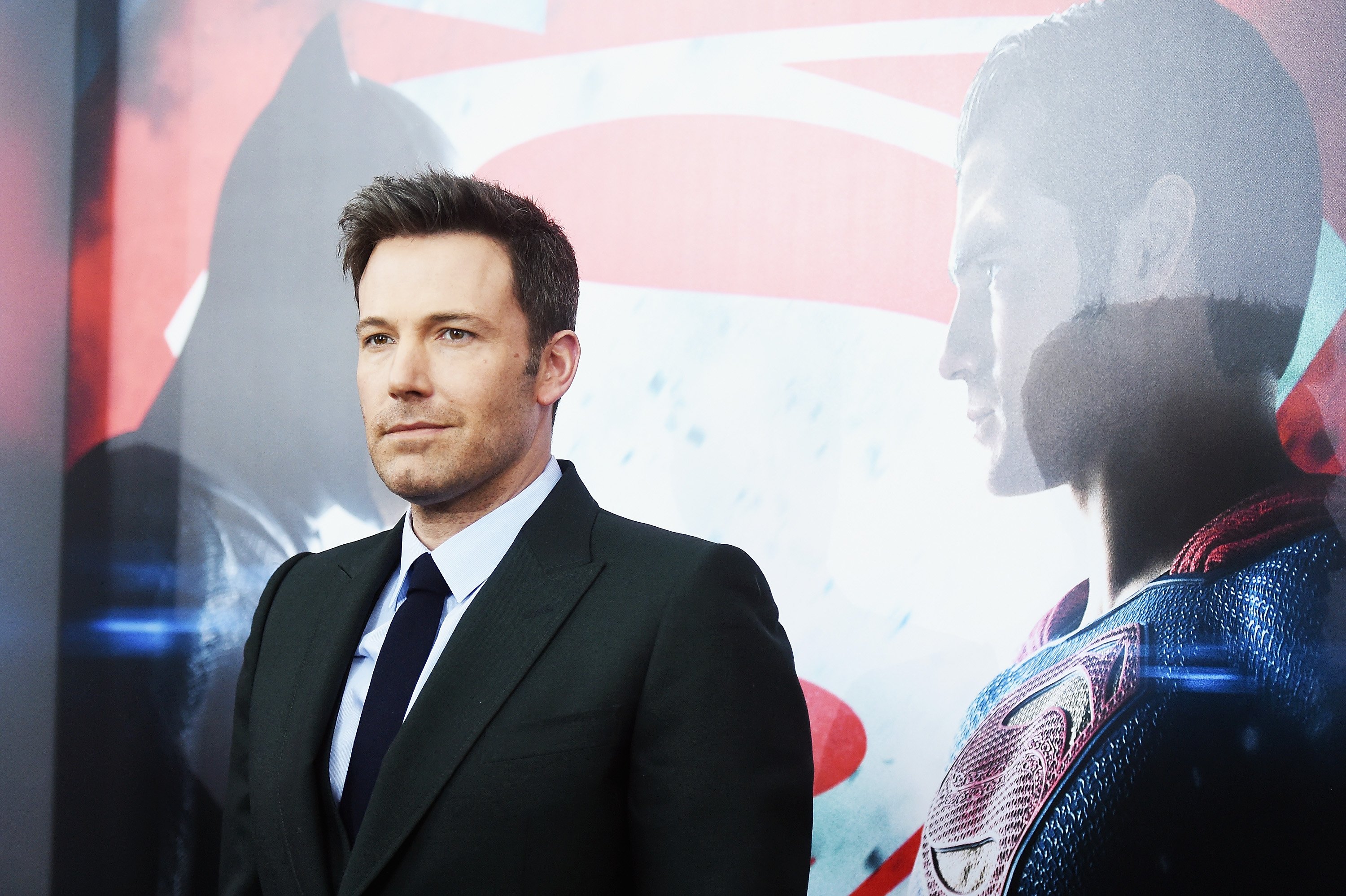 Ben Affleck is smiling in front of a Batman V Superman: Dawn Of Justice poster.