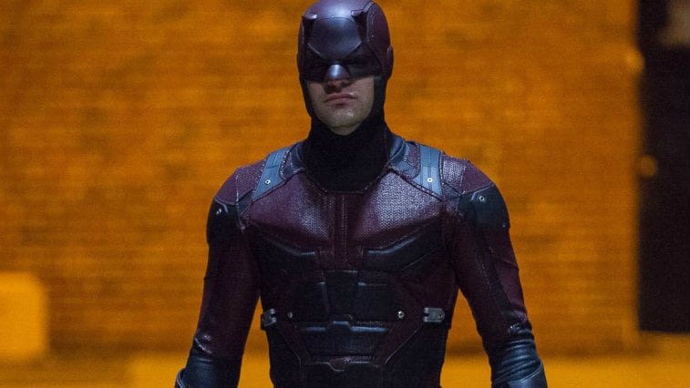 Charlie Cox wearing the full Daredevil outfit, against an orange-lit brick wall