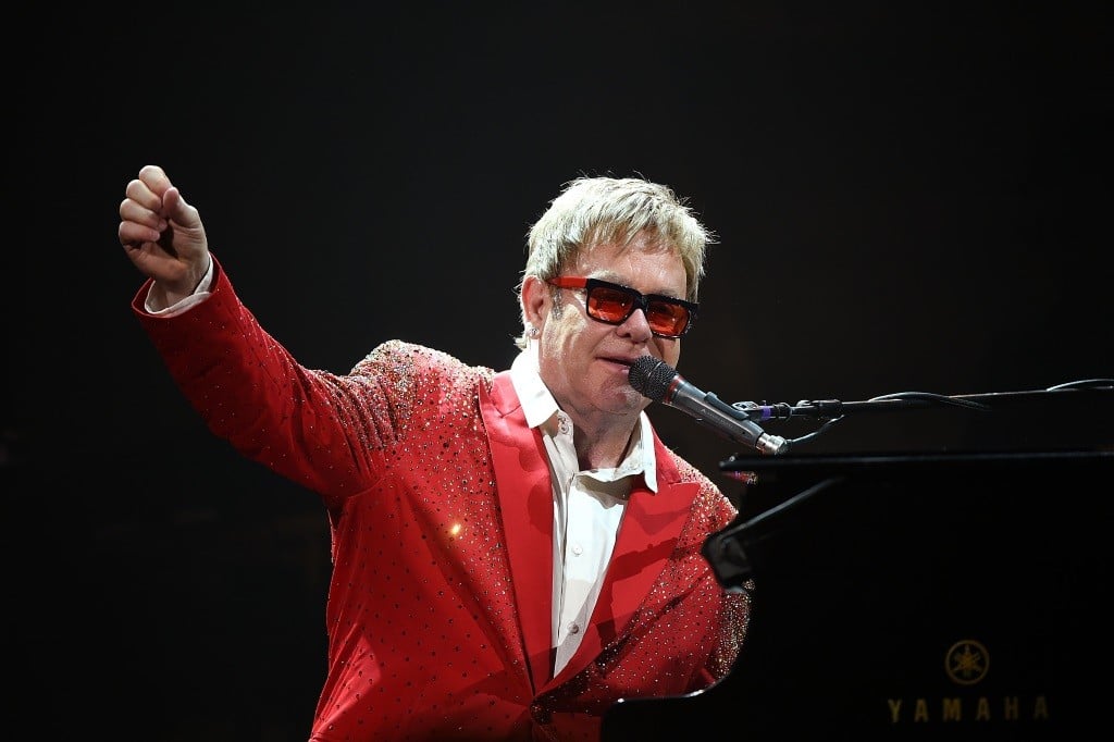 Elton John performing and playing piano, with his right arm out