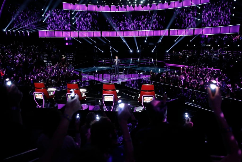Carson Daly stands on the stage of the voice in front of the four coach chairs.