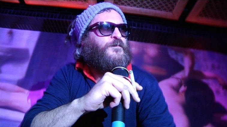 Joaquin Phoenix wearing sunglasses and a knit hat holding a microphone