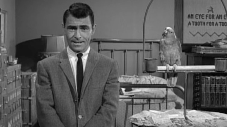 Rod Serling in black and white standing in front of a bed in The Twilight Zone