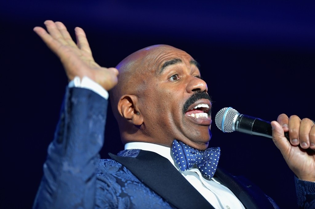 Steve Harvey holds his hands up and speaks into a microphone