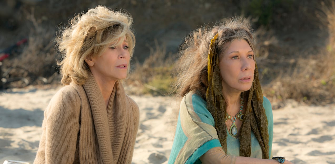 Grace and Frankie sit on a beach looking shocked