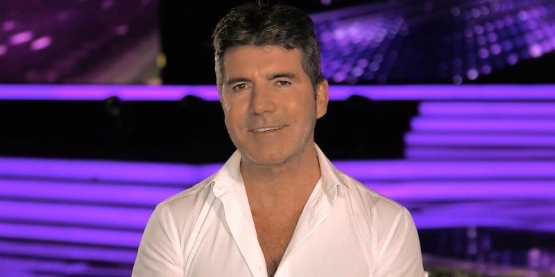 Simon Cowell is smiling in a white shirt in front of the stage of America's Got Talent.