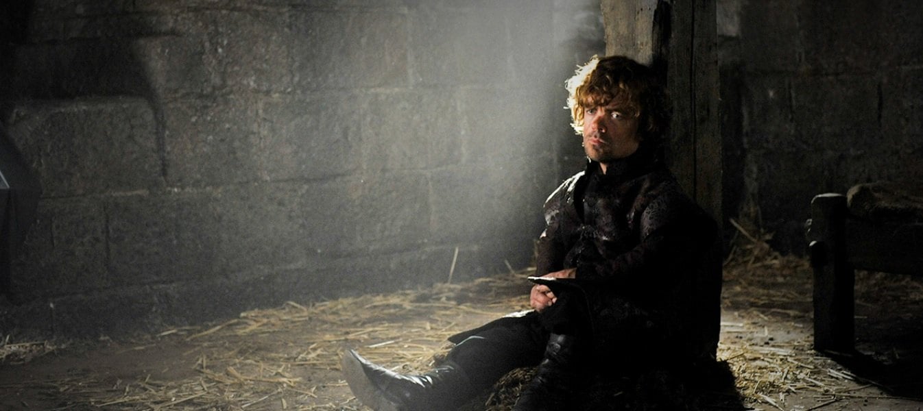 Tyrion sits up against a pillar in chains, looking down on the crowd while deeply contemplating