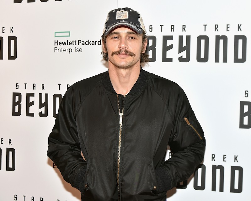 James Franco smiles while taking photos in a black jacket and cap. 