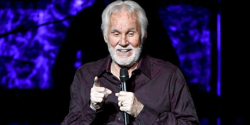 Kenny Rogers is speaking on stage holding a microphone.