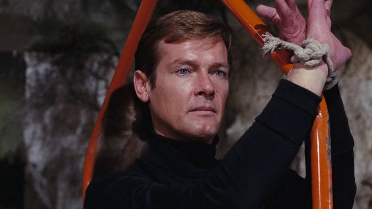 Roger Moore stares ahead as his hands are tied to a metal bar in Live and Let Die 
