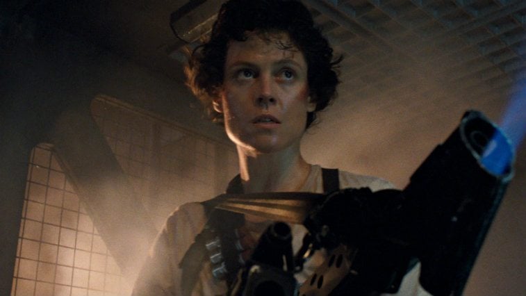 Sigourney Weaver in Aliens stands looking serious holding a machien
