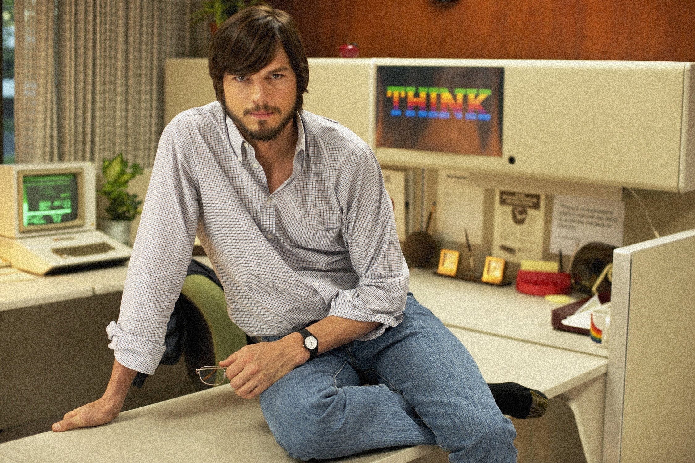 Ashton Kutcher dressed as Steve Jobs sitting on a desk.