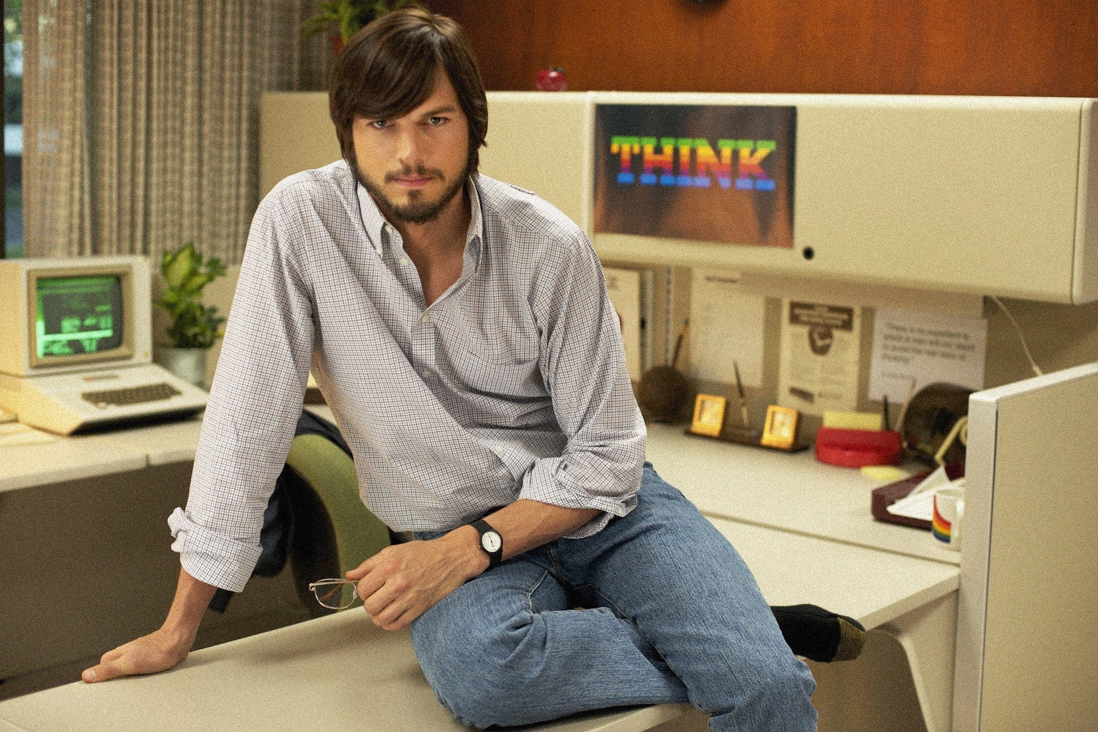 Ashton Kutcher as Steve Jobs sitting on a desk in front of an old computer and a 'THINK' poster in Jobs the film