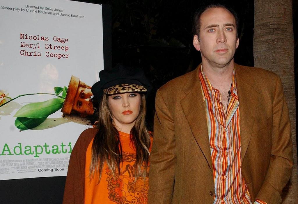 Lisa Marie Presley and Nicolas Cage looking at the camera, standing next to a poster for Adaptation