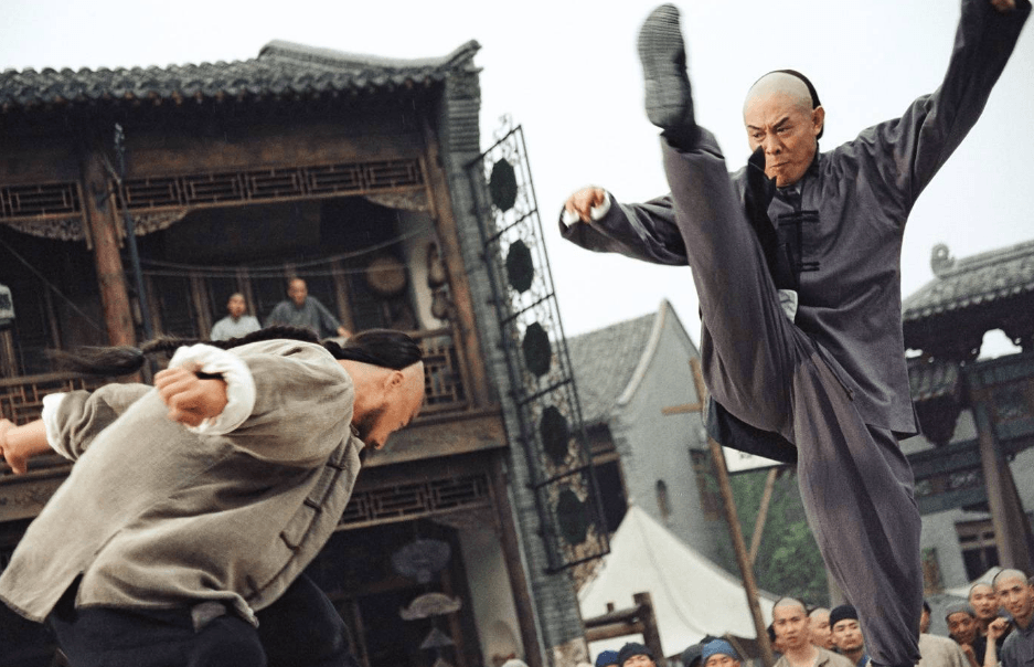 Jet Li kicks his right leg all the way up, jumping at an assailant bending over with both his arms out