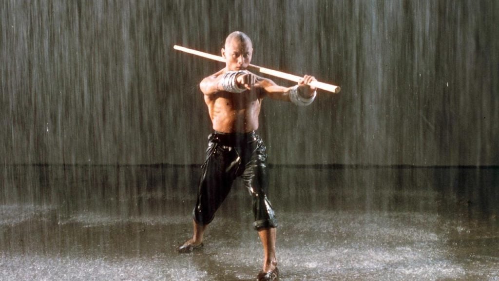 A shirtless man in the rain, holding a long staff over his shoulder, with his other fist pointed out