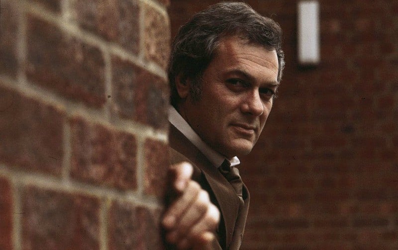 Tony Curtis peeks out from behind a brick wall while wearing a brown suit