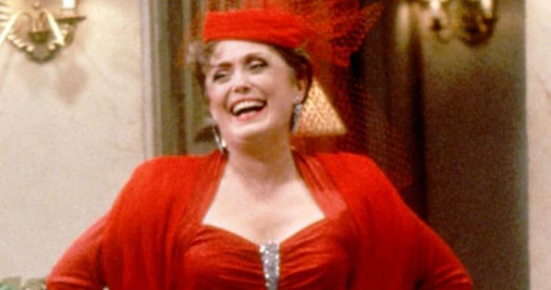 Rue McClanahan laughs in a bright red dress and hat while on the set of The Golden Girls