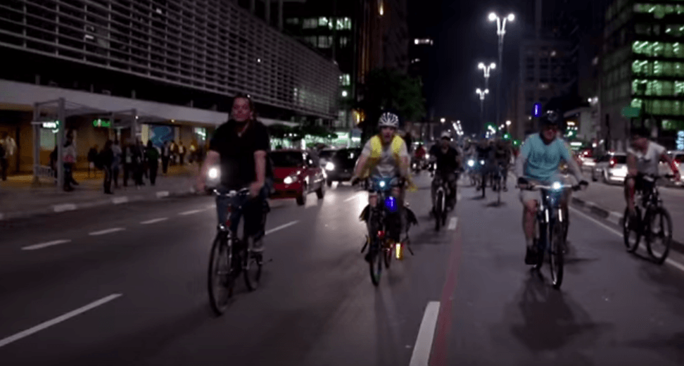 Many people in bikes riding down a street at night alongside cars in Bikes vs. Cars