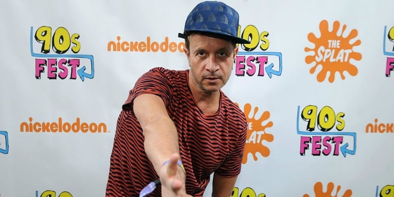 Pauly Shore is posing on the red carpet and is wearing a striped shirt and a hat.