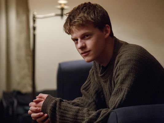Patrick sits on the couch with his hands folded in a scene from 'Manchester By the Sea.'