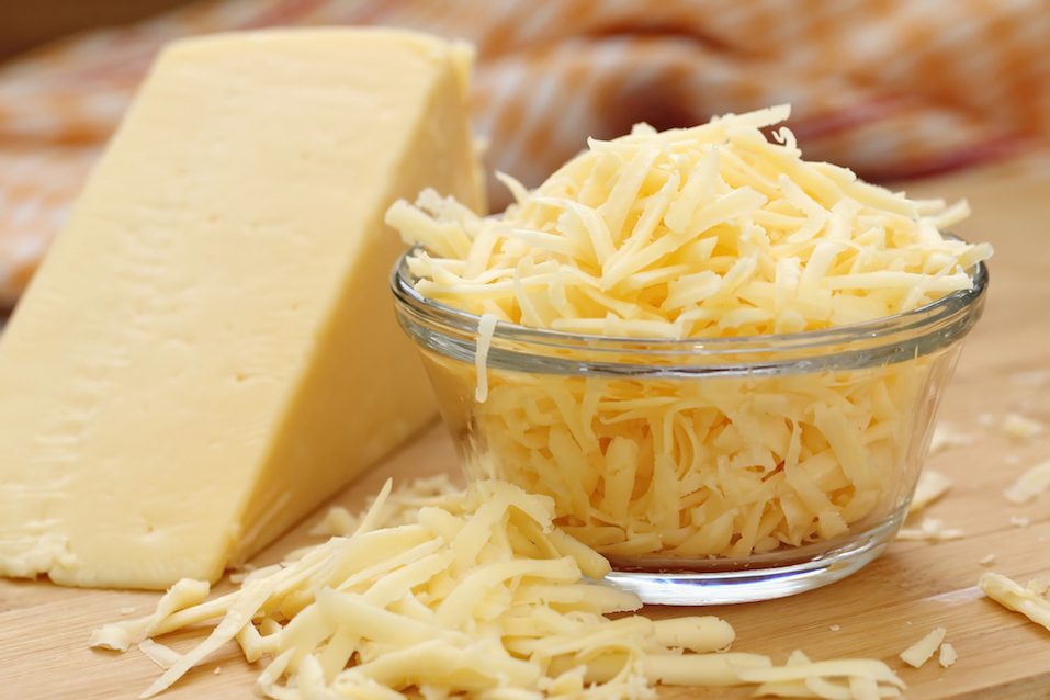 Glass bowl filled with grated cheese next to a wedge of cheese on a table.
