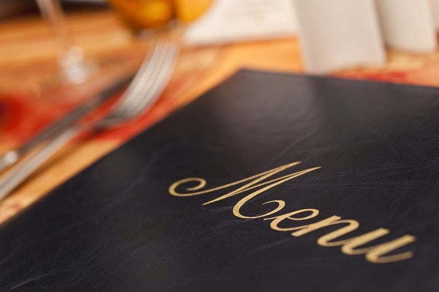 A menu resting on a restaurant table, emphasis placed on the golden embroidery lettering against the black with the image's focus. Silverware, napkins and glasses sit in the background as blurry shapes at the menu's edge, all resting on a light brown table.