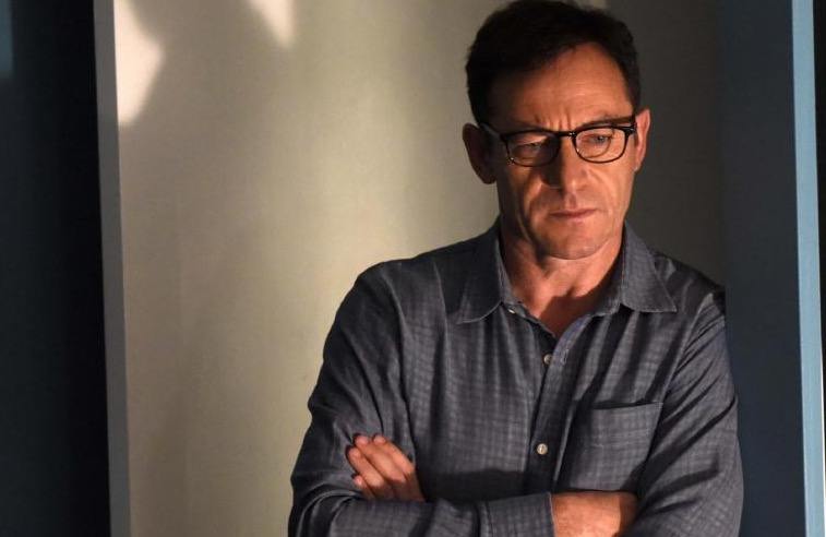 Jason Isaacs wearing glasses with his arms folded, leaning against a wall and looking down