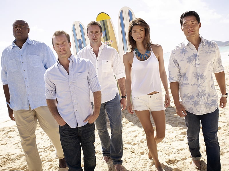 The cast of Hawaii Five-O walks on a beach with surfboards and the ocean in the background