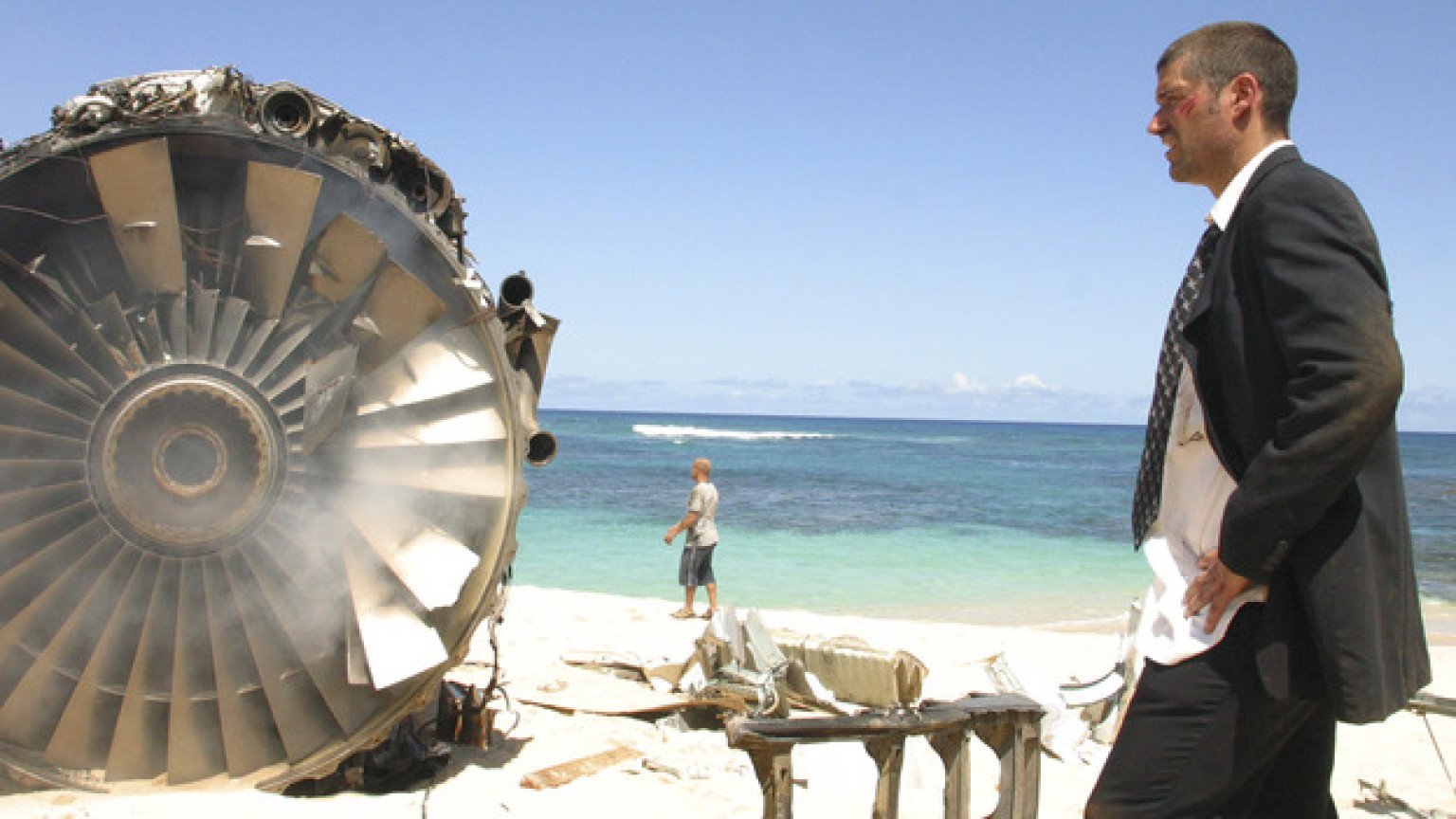 Matthew Fox in a suit, standing next to a plane engine on a beach