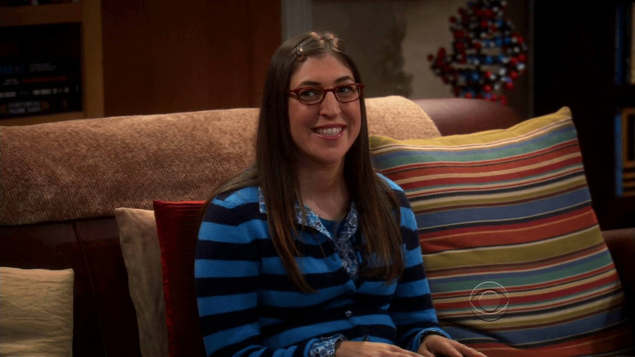 Mayim Bialick, wearing a blue striped sweater, and smiling on a couch