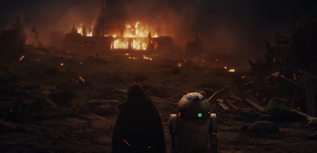 Luke and R2D2 look on at a house on fire, amidst a ruin