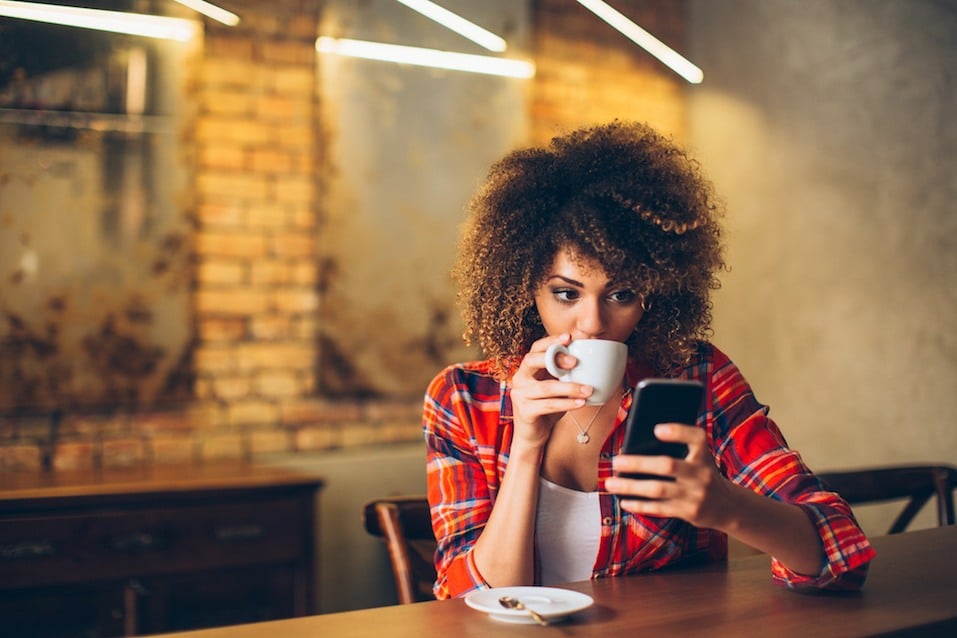 Young woman drinking coffee and researching flights on her smartphone