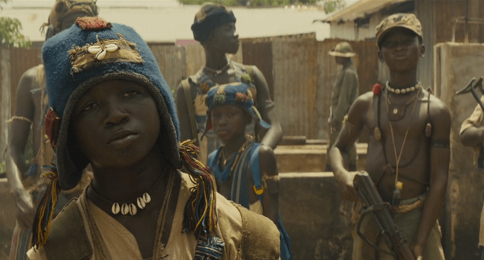 Agu stands looking defiant as child soldiers stand behind him holding weapons in a scene from 'Beasts of No Nation.'