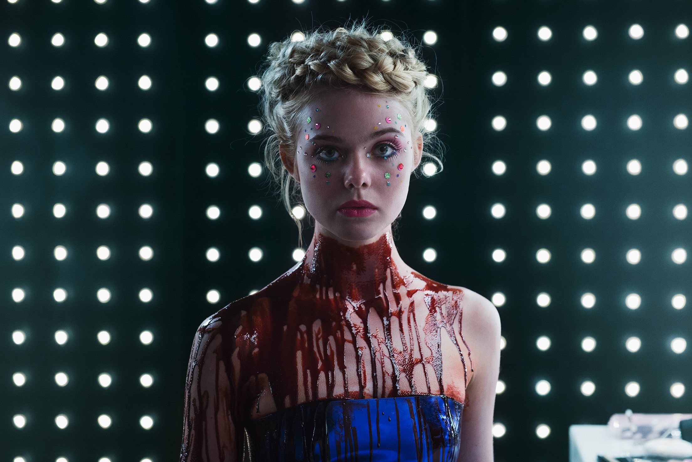 Jesse (Elle Fanning) stands looking at the camera, her face decorated with jewels, and blood covering her neck and chest, in 'Neon Demon.'